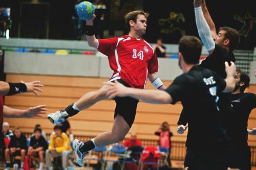 Handball: Spieler im roten Trikot beim Sprungwurf vor der Abwehr